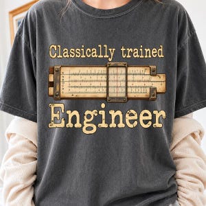 Puede incluir: Una camiseta gris carbón con un gráfico de una regla de cálculo de madera con un cursor central. El texto "Classically trained" aparece sobre la regla y la palabra "Engineer" está impresa debajo en letras grandes de color beige desgastado.