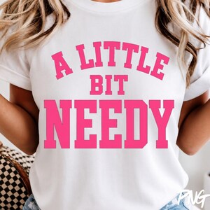 以下が含まれることがあります： 胸元に鮮やかなピンク色のカレッジ風フォントで「A LITTLE BIT NEEDY」というテキストがプリントされた、白い半袖Tシャツ。クルーネックで標準的なフィット感のデザインとなっており、グラフィックは前面の中央に配置されています。
