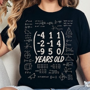May include: A black short-sleeved t-shirt featuring a white graphic print of a mathematical matrix with the numbers -4, 1, 1, -2, -1, 4, -9, 5, 0 followed by the text YEARS OLD, surrounded by various white mathematical formulas and geometric shapes.
