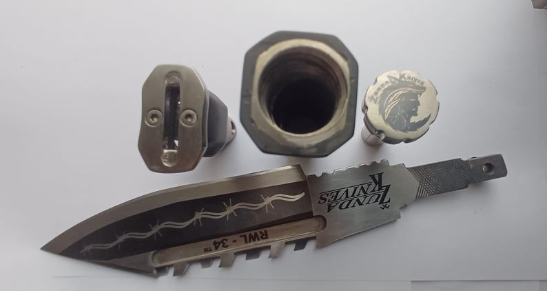 May include: A silver knife with a black handle and a barbed wire design. The knife has a black and silver blade with the text "ZUNDA KNIVES" and "RWL - 34" engraved on it. The knife also has a silver cap with a design of a person's head and the text "ZUNDA KNIVES" engraved on it.
