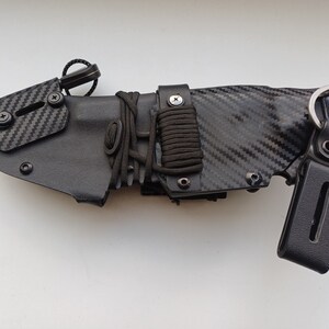 May include: A black knife sheath with a carbon fiber pattern and a black strap with a metal buckle. The sheath has a black cord wrapped around it and a black metal loop.