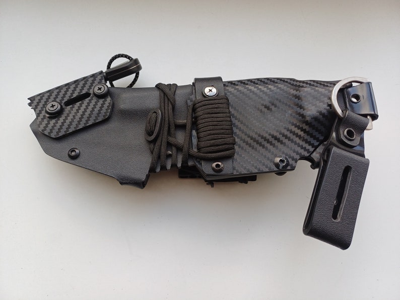 May include: A black knife sheath with a carbon fiber pattern and a black strap with a metal buckle. The sheath has a black cord wrapped around it and a black metal loop.