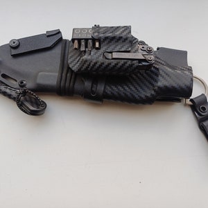 May include: A black knife sheath with a carbon fiber pattern and a multi-tool attached to it. The sheath has a small loop on the side and a key ring attached to the other side.