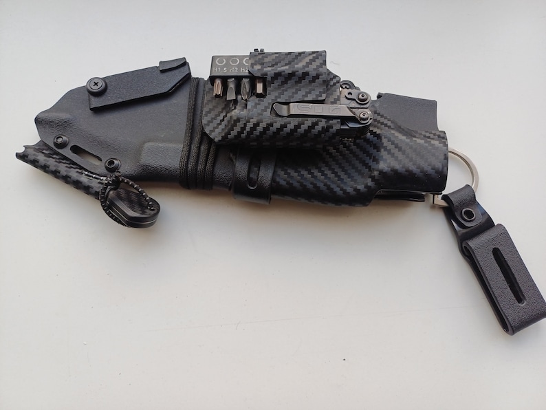 May include: A black knife sheath with a carbon fiber pattern and a multi-tool attached to it. The sheath has a small loop on the side and a key ring attached to the other side.