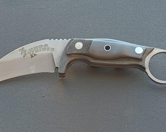 May include: A curved karambit knife with a silver blade and a dark handle. The blade has the text "ZANIVES" etched on it. The handle features a finger ring and a textured grip for secure handling. The knife is designed for close-quarters combat.