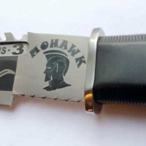 May include: A black and silver folding knife with a serrated blade. The knife has a black handle and a silver blade with the words "Survival Systems - 3" and "Mohawk" engraved on it.