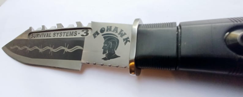May include: A black and silver folding knife with a serrated blade. The knife has a black handle and a silver blade with the words "Survival Systems - 3" and "Mohawk" engraved on it.