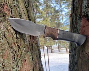 May include: A handcrafted hunting knife with a dark, textured blade and a brown and black handle. The knife is positioned between two tree trunks, with a snowy landscape in the background. The blade appears to be approximately 8 inches long.
