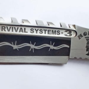 May include: A silver knife with a black handle and a black blade with a barbed wire design. The blade is engraved with the text "SURVIVAL SYSTEMS - 3" and "MOHAWK".
