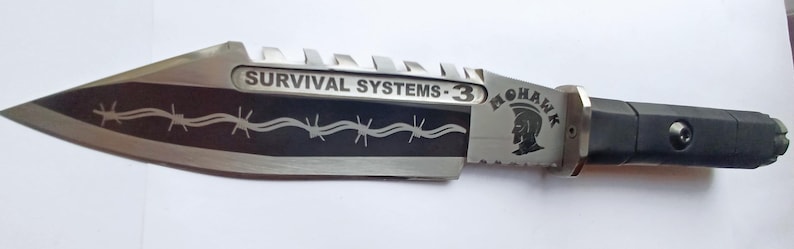 May include: A silver knife with a black handle and a black blade with a barbed wire design. The blade is engraved with the text "SURVIVAL SYSTEMS - 3" and "MOHAWK".