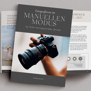 Op de afbeelding: Een zwarte en grijze boekomslag met de titel "Fotografieren im Manuellen Modus" in witte tekst. De ondertitel is "Sag "Tschüss" zum Programm-Modus - für immer." De omslag toont ook een foto van een persoon die een zwarte camera lens vasthoudt.
