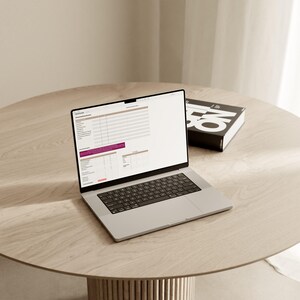 May include: A silver laptop computer with a black keyboard is open on a light brown wooden table. The laptop screen displays a spreadsheet with data and charts. A black book with white text is on the table to the right of the laptop.