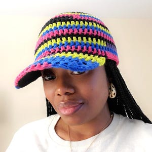 Puede incluir: Gorra de béisbol de ganchillo multicolor con visera curva. La gorra presenta rayas horizontales negras, rosas, amarillas y azules. La visera es negra por debajo. La persona de la imagen lleva una camiseta blanca con un estampado gráfico.
