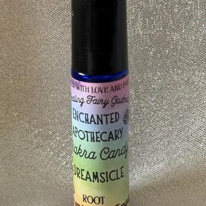 Puede incluir: Una pequeña botella cilíndrica de vidrio con tapa negra. La botella tiene una etiqueta con las palabras "ENCHANTED APOTHECARY Chakra Candy DREAMSICLE" en un degradado arcoíris. También es visible el texto "ROOT".