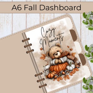 Könnte beinhalten: Ein A6-Planer-Dashboard mit einem braunen Teddybären, der einen Schal trägt und hinter einem großen orangefarbenen Kürbis sitzt. Der Text Cozy Moments ist in schwarzer Schreibschrift über dem Bären gedruckt. Das Design zeigt Herbstblätter und Eicheln.