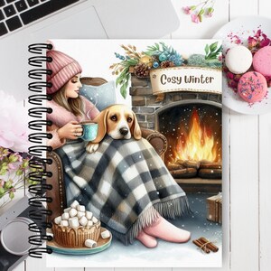 May include: Spiral-bound notebook with a cozy winter scene. The illustration depicts a person and a dog relaxing by a fireplace. The scene includes a blanket, a cup of coffee, and the words "Cosy Winter". A plate of macarons and a cup of hot chocolate are also visible.