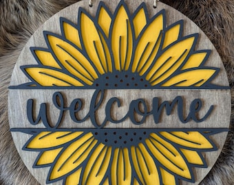 May include: A round wooden welcome sign with a black and yellow sunflower design. The word "welcome" is cut out of the wood and painted black.