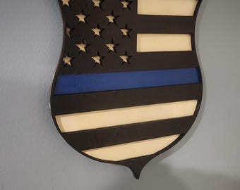 May include: A wooden shield-shaped sign with a thin blue line and an American flag design.