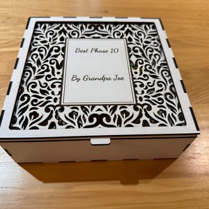 Pode incluir: Uma caixa de madeira branca com um design decorativo vazado na tampa. A tampa apresenta o texto "Best Phase 10 By Grandpa Joe" em uma moldura retangular. A caixa está fechada e repousa sobre uma superfície de madeira.