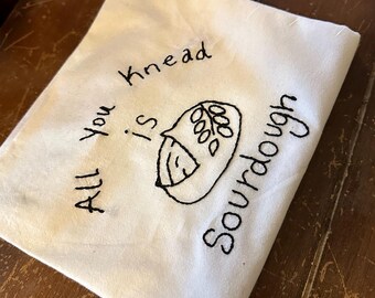 Op de afbeelding: Een witte keukendoek met zwarte borduurwerk dat "Alles wat je nodig hebt is zuurdesem" zegt. Het borduurwerk bevat een eenvoudige tekening van een zuurdesembrood.