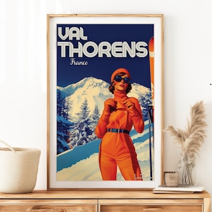 May include: Framed vintage travel poster for Val Thorens, France. The artwork features a woman in an orange ski suit and sunglasses, standing in front of a snowy mountain scene. The poster has the text "VAL THORENS France" at the top.