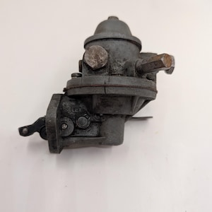 May include: A vintage, grey Solex fuel pump. The pump has a domed top, multiple bolts, and a lever on the side. The word "SOLEX" is visible on the side of the pump. The pump appears to be made of metal and shows signs of age.