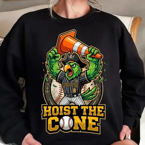 Pode incluir: Camisola preta com um papagaio de desenho animado usando um chapéu de pirata e uniforme de beisebol, segurando um cone de trânsito laranja. O design inclui o texto "HOIST THE CONE" e uma bola de beisebol.