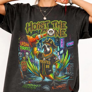 K&ouml;nnte beinhalten: Schwarzes T-Shirt mit einem bunten Papagei, der einen Piratenhut und ein Baseball-Trikot tr&auml;gt und einen orangefarbenen Verkehrskegel h&auml;lt. Der Text lautet "Hoist the Cone". Das Design beinhaltet Totenk&ouml;pfe und die Worte "Urban Pirates."