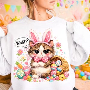 May include: White sweatshirt with a cartoon cat wearing bunny ears and a pink bow tie. The cat has an angry expression and the word "WHAT?" in a speech bubble. Easter eggs and a basket are also in the design.