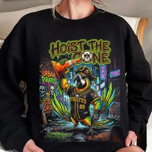 K&ouml;nnte beinhalten: Schwarzes Sweatshirt mit einem farbenfrohen Papageien-Motiv. Der Papagei tr&auml;gt einen Piratenhut und ein Trikot mit der Aufschrift "Pirates 89" und h&auml;lt einen orangefarbenen Verkehrskegel. Der Text lautet "Hoist the Cone" und "Urban Pirates".