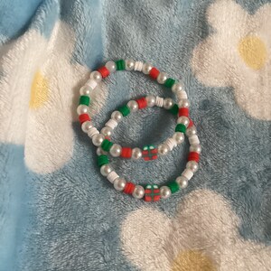 May include: Two beaded bracelets with white, red and green beads. Each bracelet has a small red and green gift box charm.