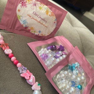 K&ouml;nnte beinhalten: Drei rosa Rei&szlig;verschlusstaschen mit wei&szlig;en Perlen- und bunten Perlenketten darin. Die Taschen haben ein wei&szlig;es ovales Etikett mit dem Text "Ella's Jewellery" und der E-Mail-Adresse "ellas_jewellery@outlook.com".