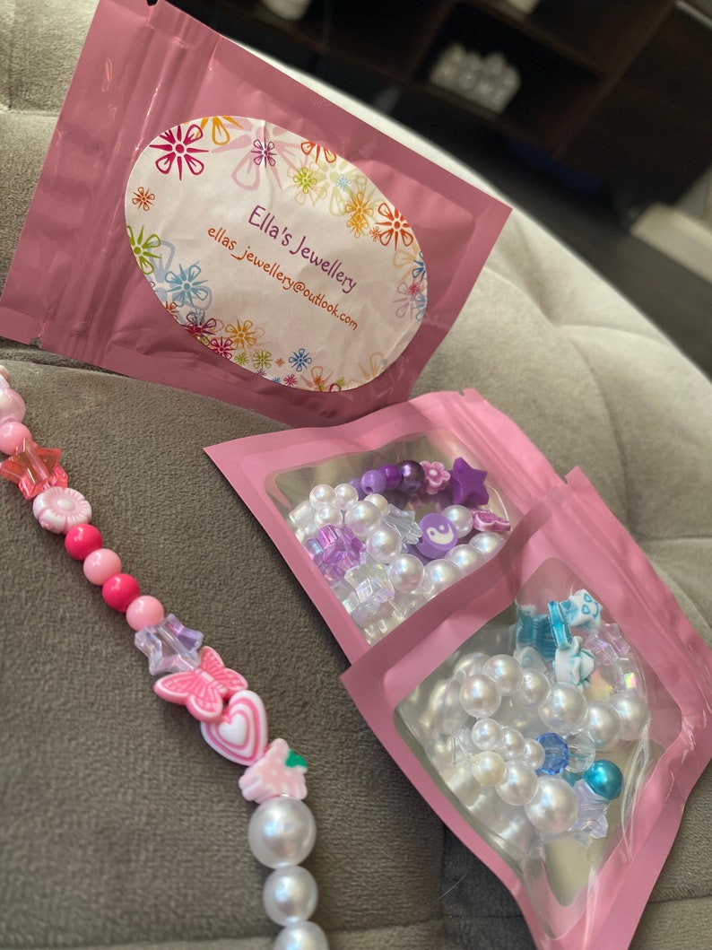 K&ouml;nnte beinhalten: Drei rosa Rei&szlig;verschlusstaschen mit wei&szlig;en Perlen- und bunten Perlenketten darin. Die Taschen haben ein wei&szlig;es ovales Etikett mit dem Text "Ella's Jewellery" und der E-Mail-Adresse "ellas_jewellery@outlook.com".