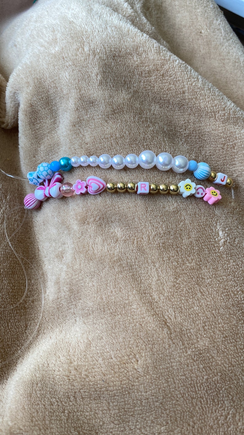 K&ouml;nnte beinhalten: Eine Perlenkette mit wei&szlig;en Perlen, goldenen Perlen und bunten Charms, darunter ein Herz, eine Blume, ein Smiley und die Buchstaben "R" und "J".