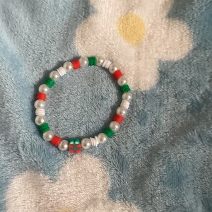 May include: A white, red and green beaded bracelet with a small gift box charm. The bracelet is on a blue fluffy surface.