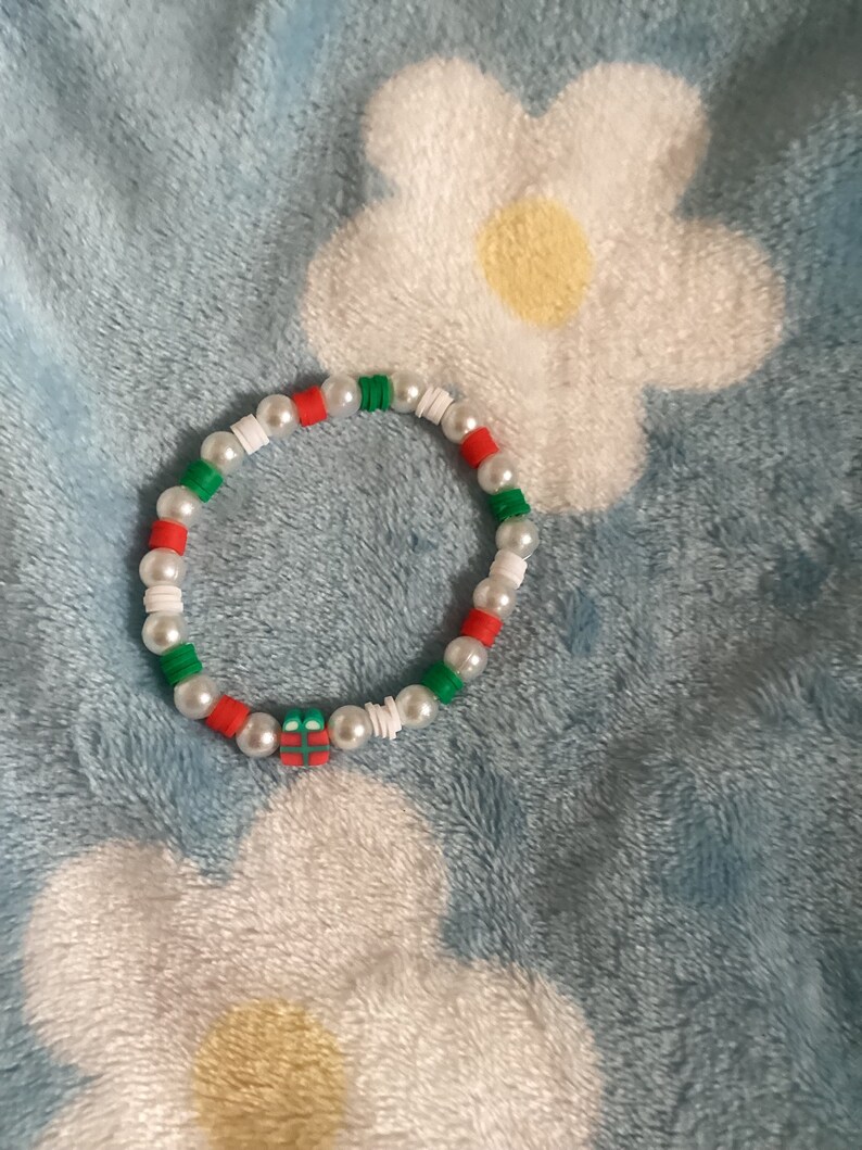 May include: A white, red and green beaded bracelet with a small gift box charm. The bracelet is on a blue fluffy surface.