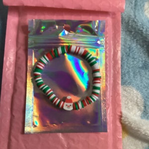 Puede incluir: Un brazalete de Navidad rojo, blanco y verde con un dije de mu&ntilde;eco de nieve. El brazalete est&aacute; hecho de cuentas de colores y est&aacute; empaquetado en una bolsa de pl&aacute;stico transparente.