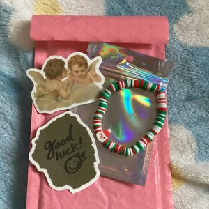 Puede incluir: Un sobre acolchado rosa con un brazalete de cuentas blancas y verdes, una pegatina con dos &aacute;ngeles y una pegatina con el texto "&iexcl;buena suerte!" y una carita sonriente.