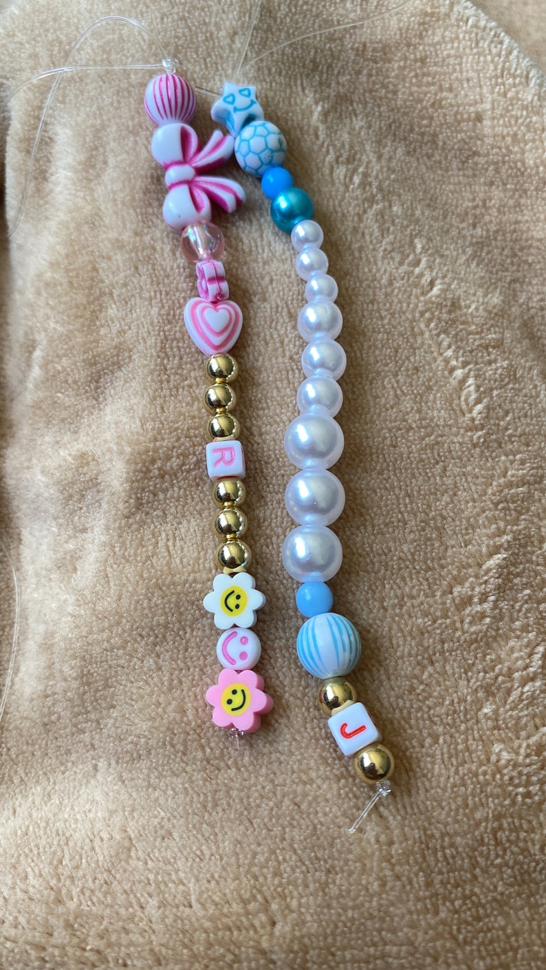 K&ouml;nnte beinhalten: Zwei Perlen-Handy-Anh&auml;nger, einer in Pink und Wei&szlig; mit einem Herz-, Blumen- und Buchstaben-Anh&auml;nger "R" und einer in Blau und Wei&szlig; mit einem Stern-, Perlen- und Buchstaben-Anh&auml;nger "J".