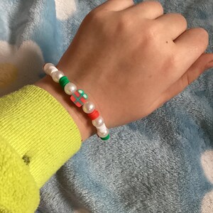 May include: A white beaded bracelet with red, green and white beads. The bracelet has a charm in the shape of a gift box with a red bow.