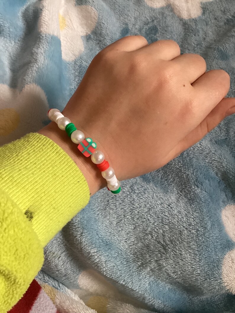 May include: A white beaded bracelet with red, green and white beads. The bracelet has a charm in the shape of a gift box with a red bow.
