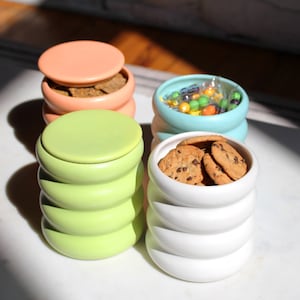 May include: Four ceramic containers with lids in different colors: light pink, light green, light blue, and white. The containers are stacked and have a wavy design. The pink container has a lid and is filled with crackers. The blue container has a lid and is filled with colorful candies. The white container has a lid and is filled with chocolate chip cookies.