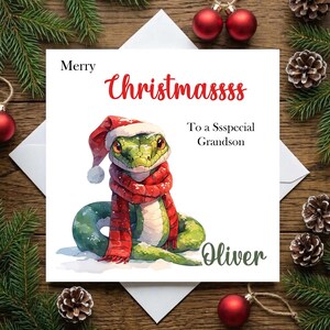May include: A square greeting card featuring a green snake wearing a red and white Santa hat and a red scarf. The text reads Merry Christmassss To a Ssspecial Grandson and the name Oliver. The design is set against a white background.