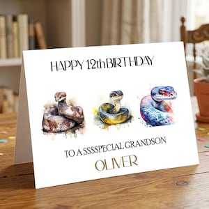 May include: A white greeting card featuring the text HAPPY 12th BIRTHDAY at the top and TO A SSSSPECIAL GRANDSON OLIVER at the bottom. The centre displays three watercolour-style illustrations of snakes in brown, yellow, and blue-purple tones.