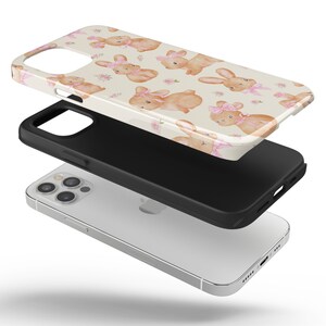 May include: A clear phone case with a design of pink-bowed bunnies on a cream background. The case is shown in three parts: the printed design, a black protective layer, and a silver phone.
