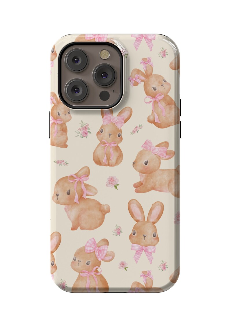 May include: A phone case with a cream background and a pattern of brown bunnies with pink bows. The case has a cutout for the camera lens.
