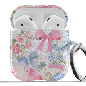 Puede incluir: Una funda blanca para Airpods con un diseño floral que presenta rosas rosas, lazos azules y un fondo blanco. La funda tiene un anillo plateado.