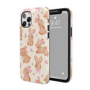 May include: A white phone case with a pattern of brown bunnies wearing pink bows and flowers. The case is designed for an iPhone with a camera bump and a clear screen protector. The phone case has a black border.
