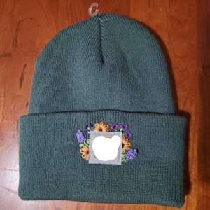 May include: A dark forest green knit beanie featuring a folded cuff. Centered on the cuff is a square patch with an embroidered floral arrangement of orange and purple flowers surrounding a white circular area in the center of the patch.