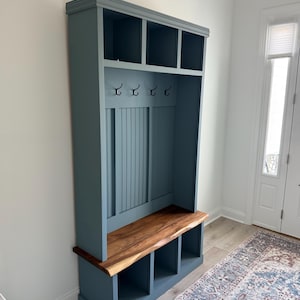 May include: A blue wooden entryway bench with a natural wood seat and storage cubbies. The bench has a built-in upper section with hooks and open shelves. The unit is painted in a muted blue color and is designed for storage and organization.